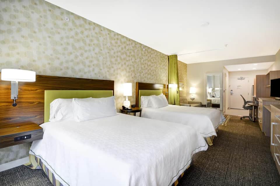 Modern hotel bedroom with two queen beds, green headboards, patterned wallpaper, and work desk