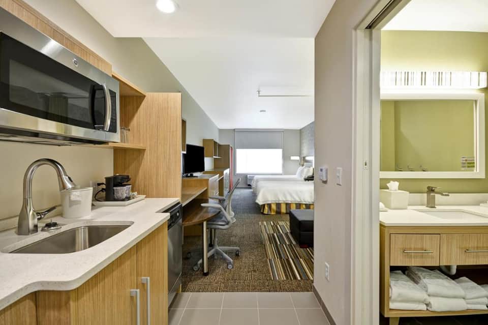 Modern hotel suite showing kitchenette, living area, bedroom with striped bedding, and bright bathroom with yellow accents