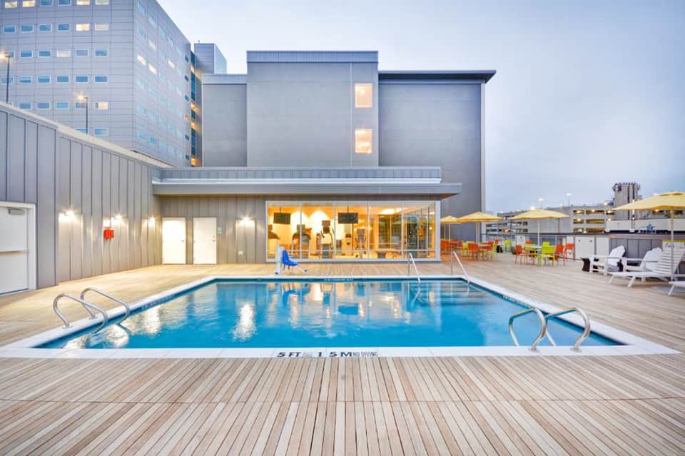 Urban rooftop pool with wooden deck overlooking city buildings at dusk with illuminated interiors