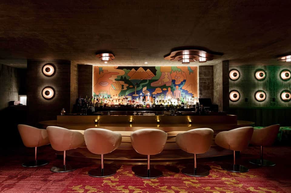 Modern hotel bar with pink upholstered chairs, colorful mural, and illuminated lighting fixtures