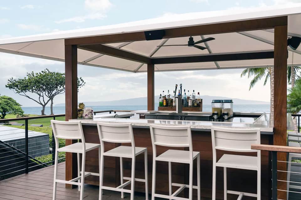 Outdoor bar with ocean view, wooden pergola, white bar stools, and retractable shade