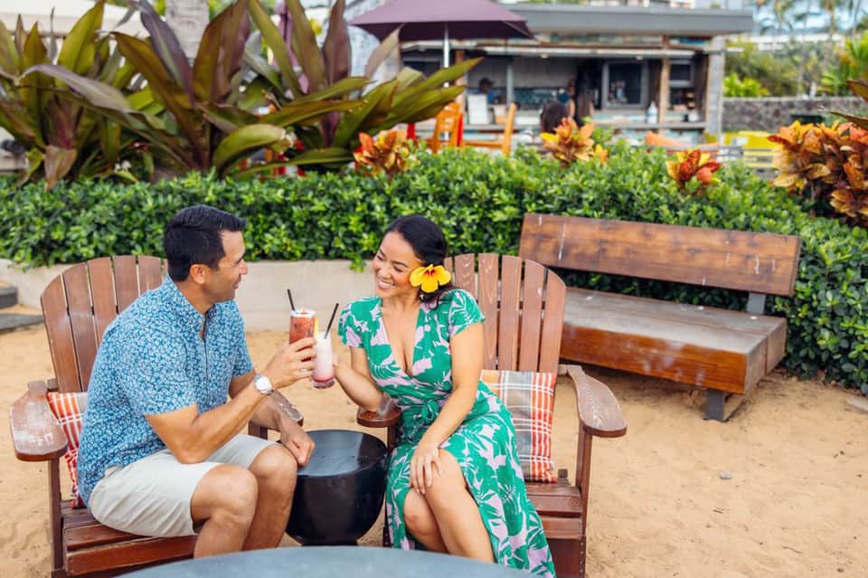 Couple toasting tropical drinks in wooden beach chairs with tropical flowers and ocean view
