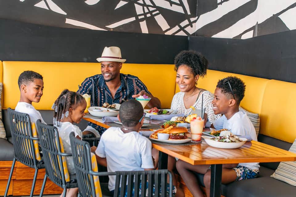 Family dining together at yellow-accented outdoor table under shade structure with tropical setting
