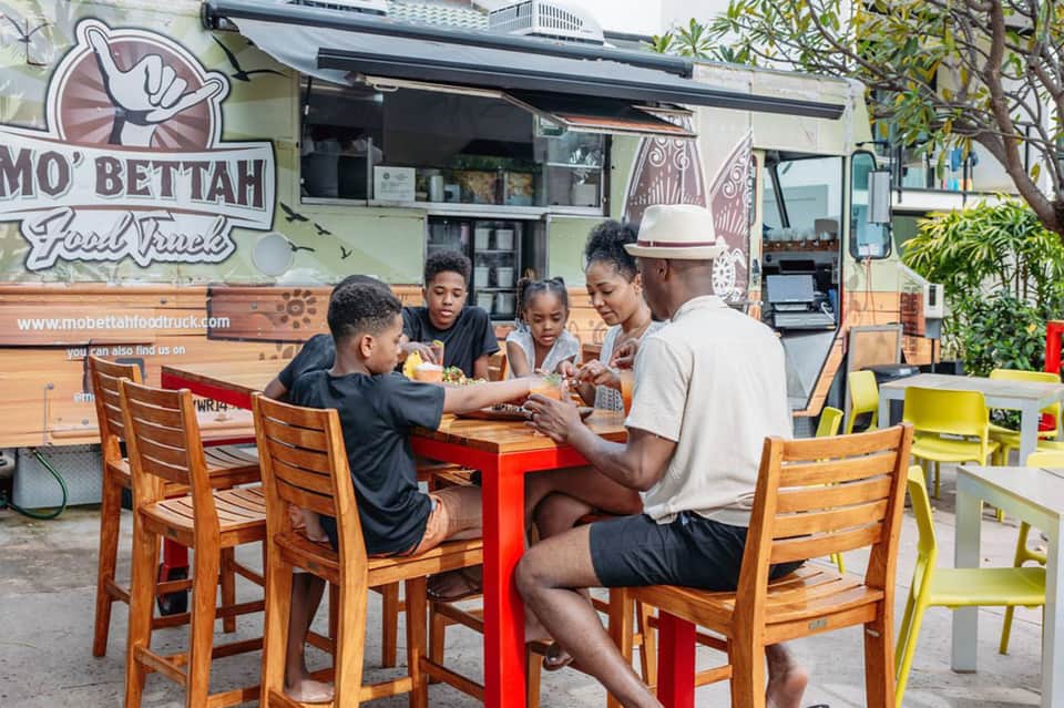 Family enjoying food truck meal outdoors at colorful tables near Mo'Bettah food truck