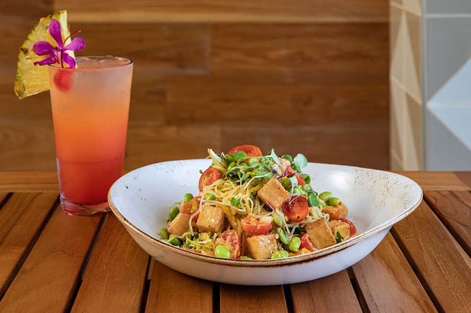 Tropical cocktail with pineapple and flower garnish beside Asian noodle bowl with tofu