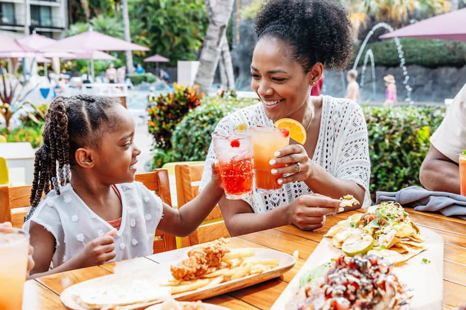 Mother and daughter enjoying tropical drinks poolside with food and palm-lined beach background