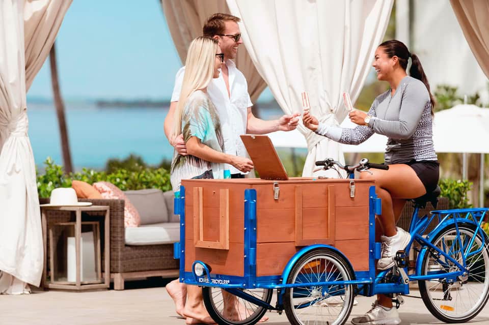 Two women at beverage cart on terrace with flowing white curtains and waterfront view