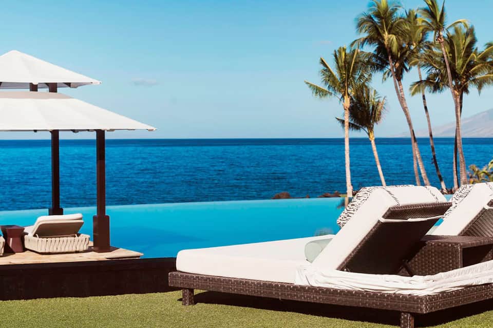 Oceanfront lounge chairs with white umbrellas overlooking infinity pool and palm trees