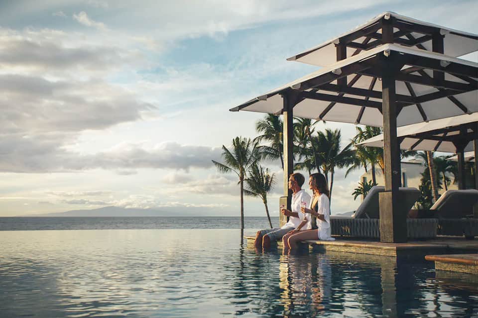 Couple relaxing under pergola overlooking infinity pool blending with ocean horizon and palm trees
