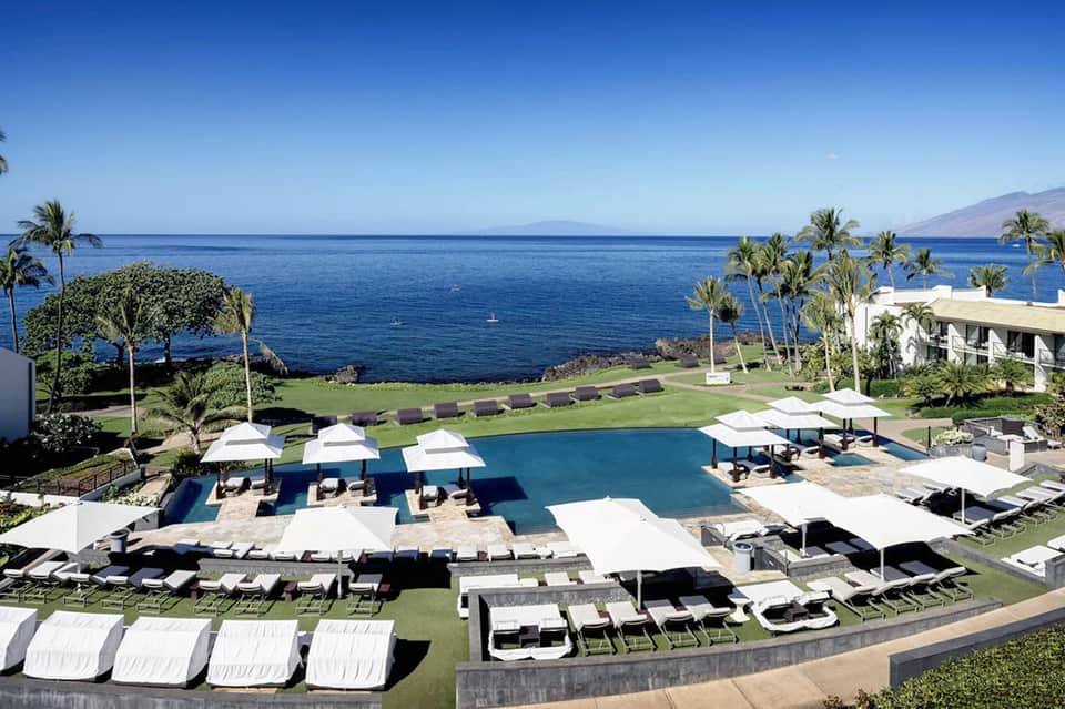 Oceanfront resort pool with white umbrellas, loungers, green lawn, and mountain seascape views