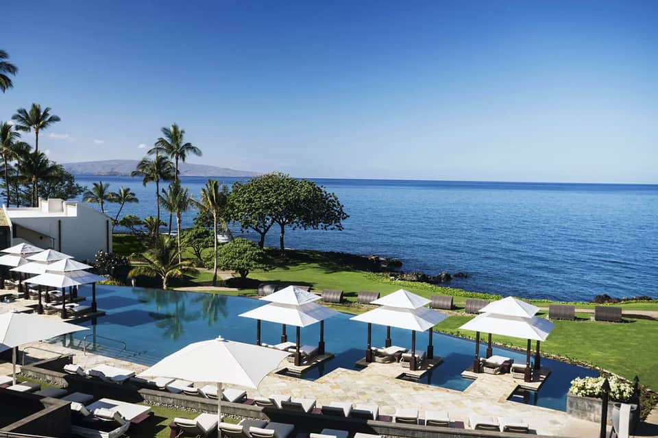 Luxury oceanfront infinity pool with white umbrellas, manicured lawn, and distant island view