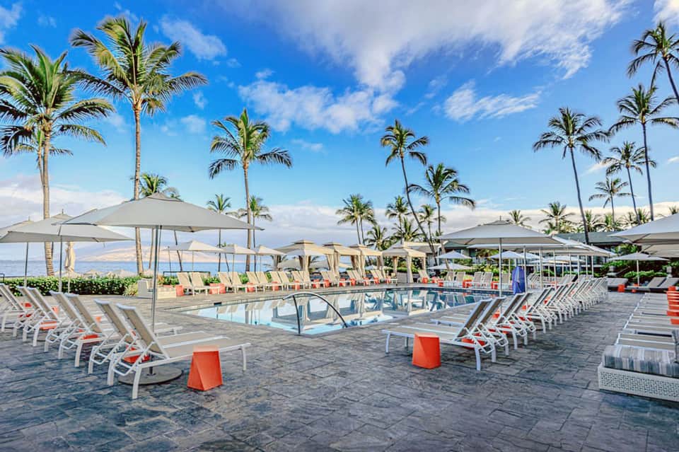 Resort pool deck with white loungers, umbrellas, palm trees, and ocean view in background.