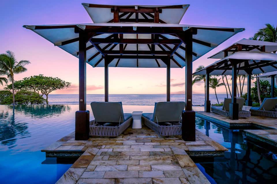 Seaside infinity pool at dusk with cabanas, palm trees, and ocean horizon