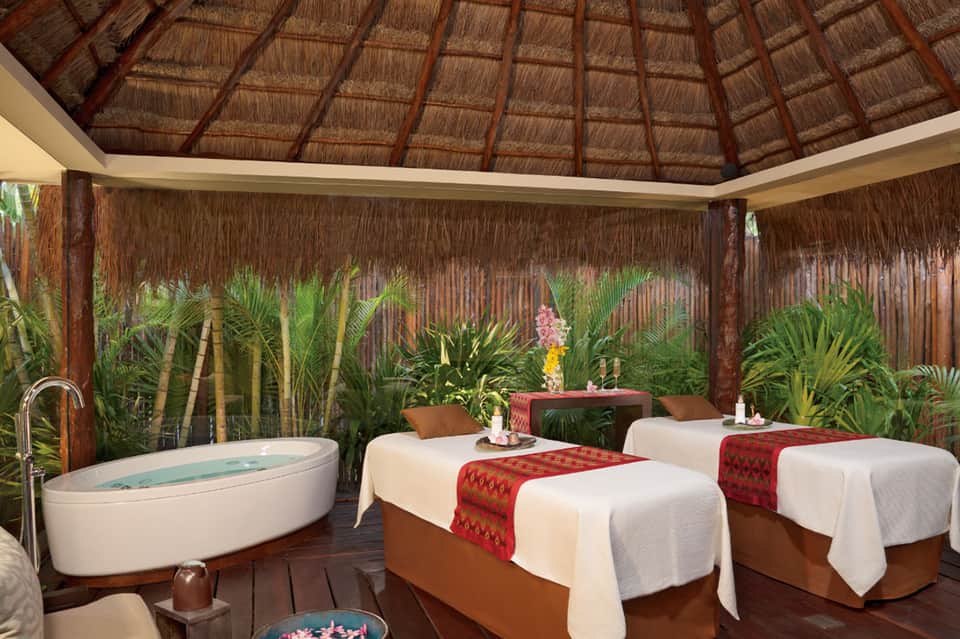 Tropical spa treatment room with thatched roof, massage beds, soaking tub, and plants