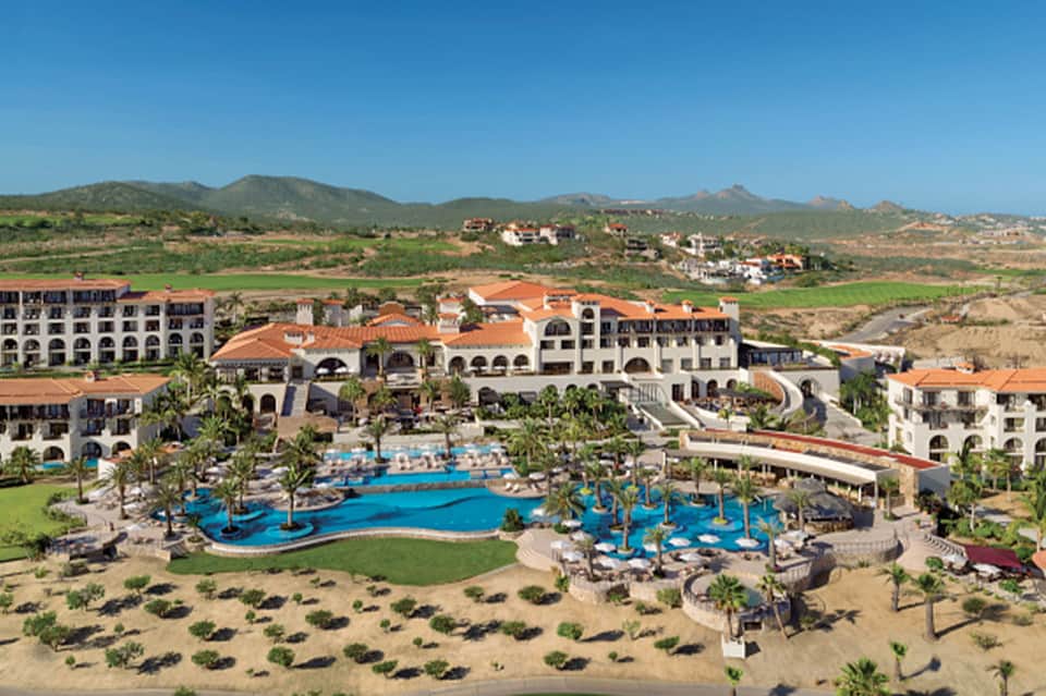 Beachfront resort with pools, palm trees, and Spanish colonial architecture against mountain backdrop