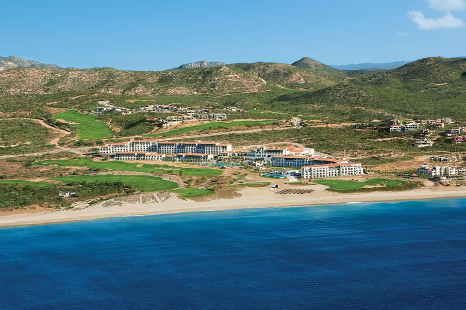 Beachfront resort nestled on hillside overlooking blue ocean, surrounded by rolling green landscape.