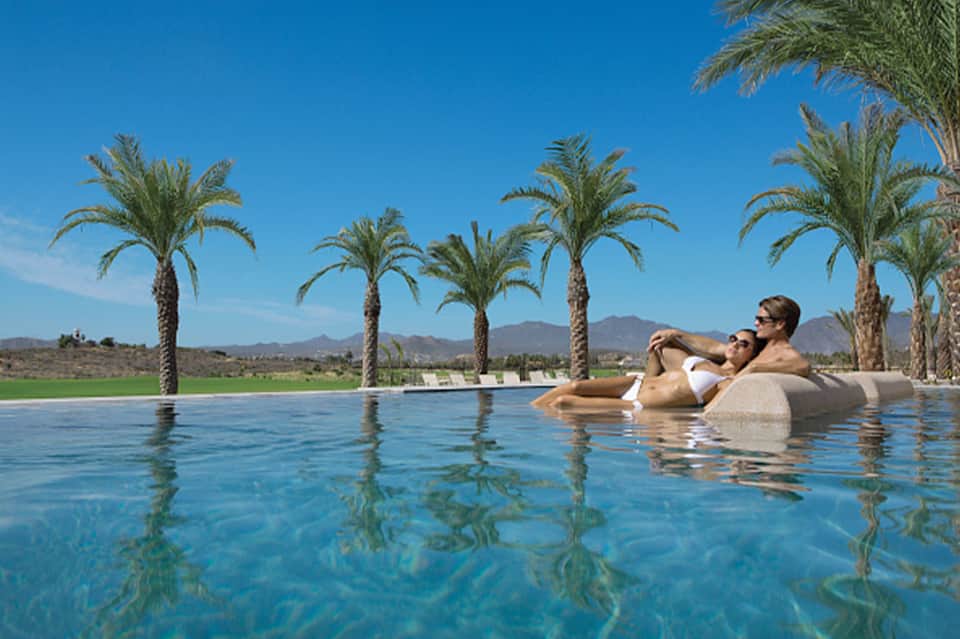 Two guests floating in infinity pool with palm trees and desert mountain landscape
