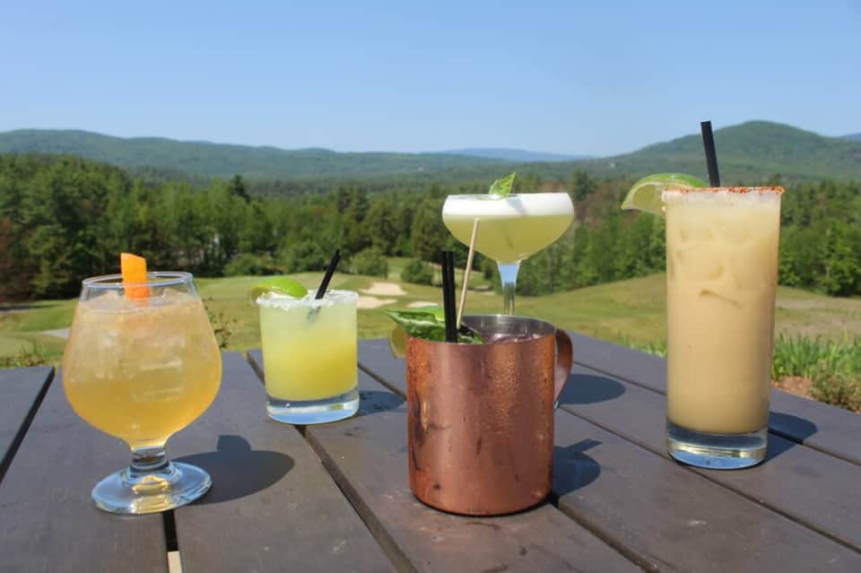 Four colorful cocktails on wooden deck overlooking forested mountain landscape