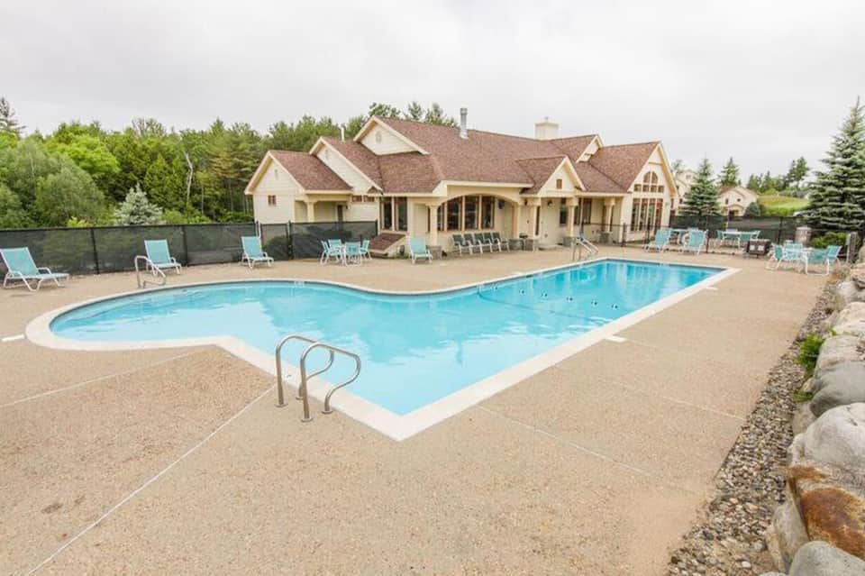 Residential backyard pool with blue water, tan concrete deck, lounge chairs, and large house in background