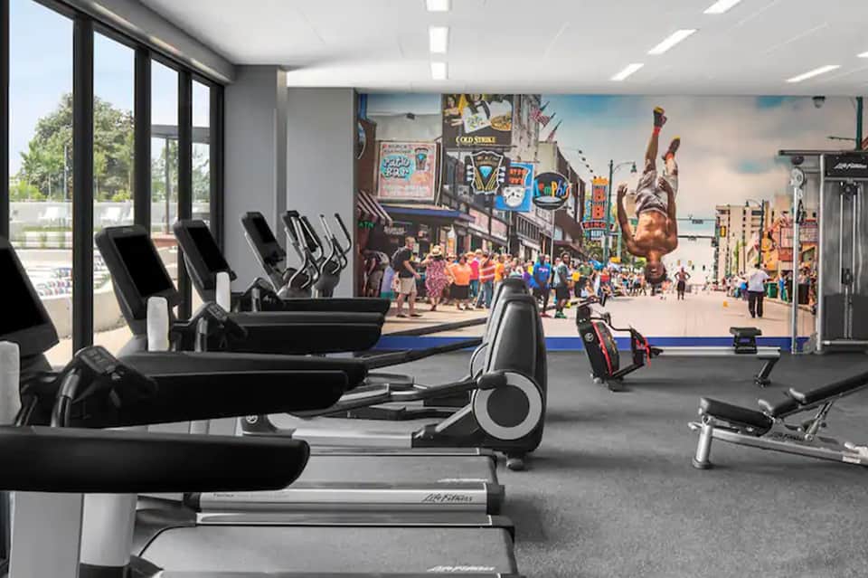 Hotel fitness center with cardio equipment, large windows, and vibrant wall mural of urban scene