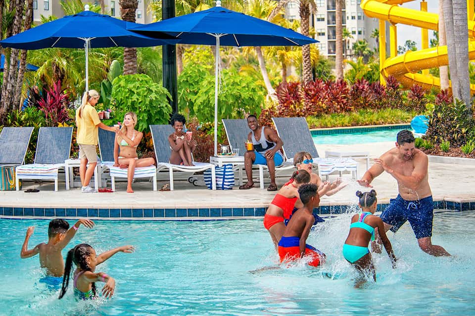 Family-friendly pool area with adults and children playing under blue umbrellas and yellow waterslide