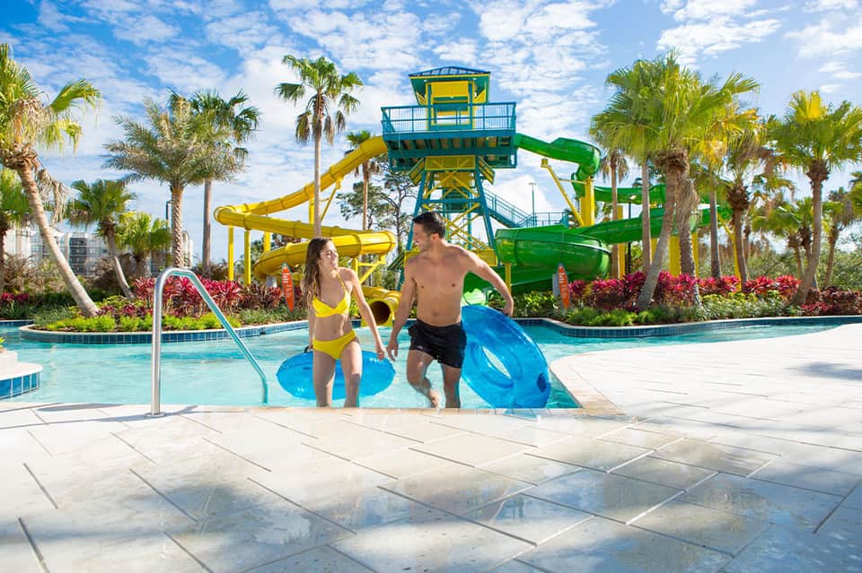 Water park with yellow and green waterslides, kids in splash pool, palm trees and sunny sky