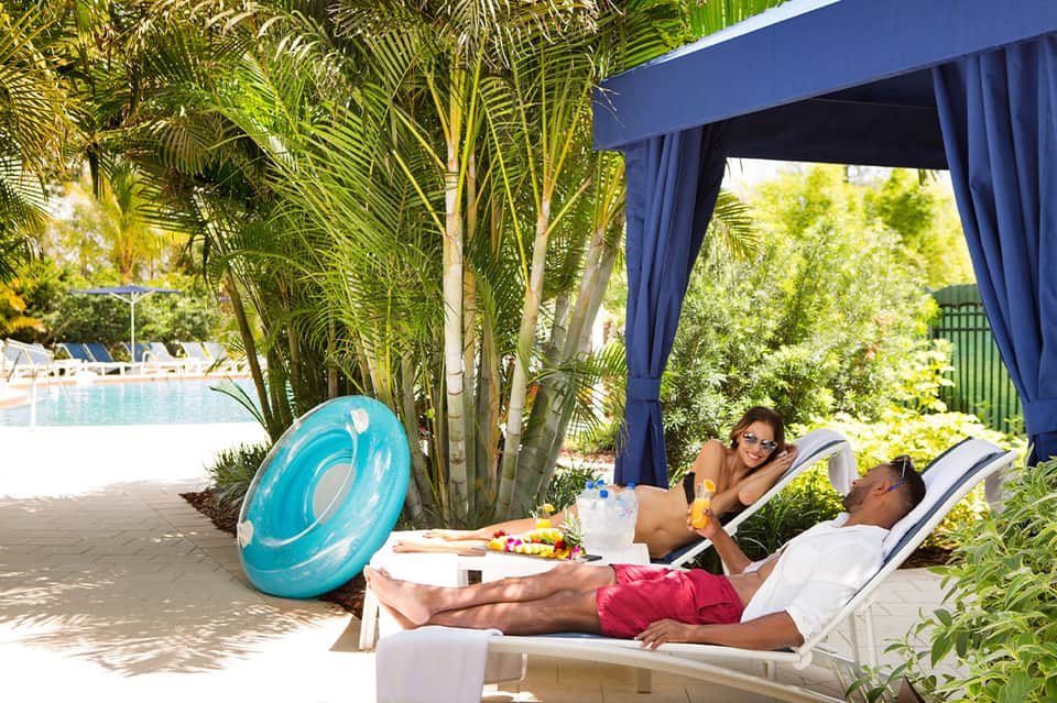 Couple relaxing on white lounge chairs under blue pergola with pool visible in background