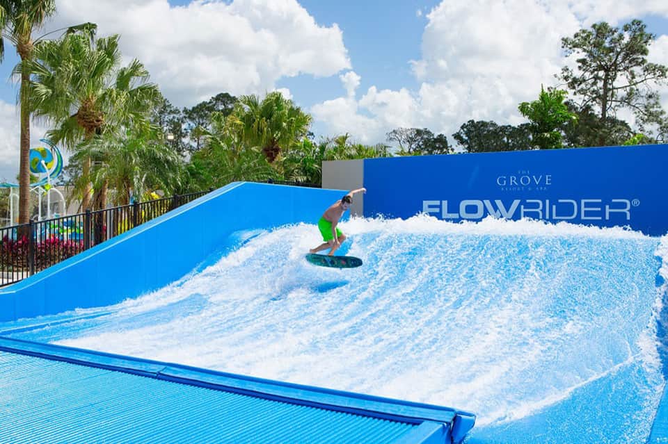 Surfer riding artificial wave at FlowRider attraction with blue water and palm trees visible