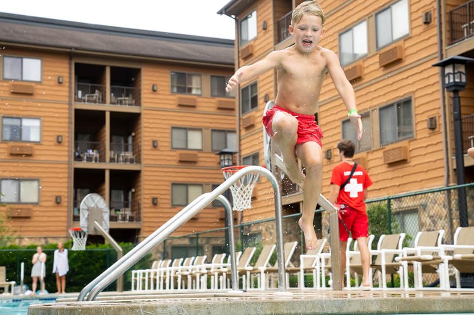 Young boy jumping from pool slide at resort with wooden building in background