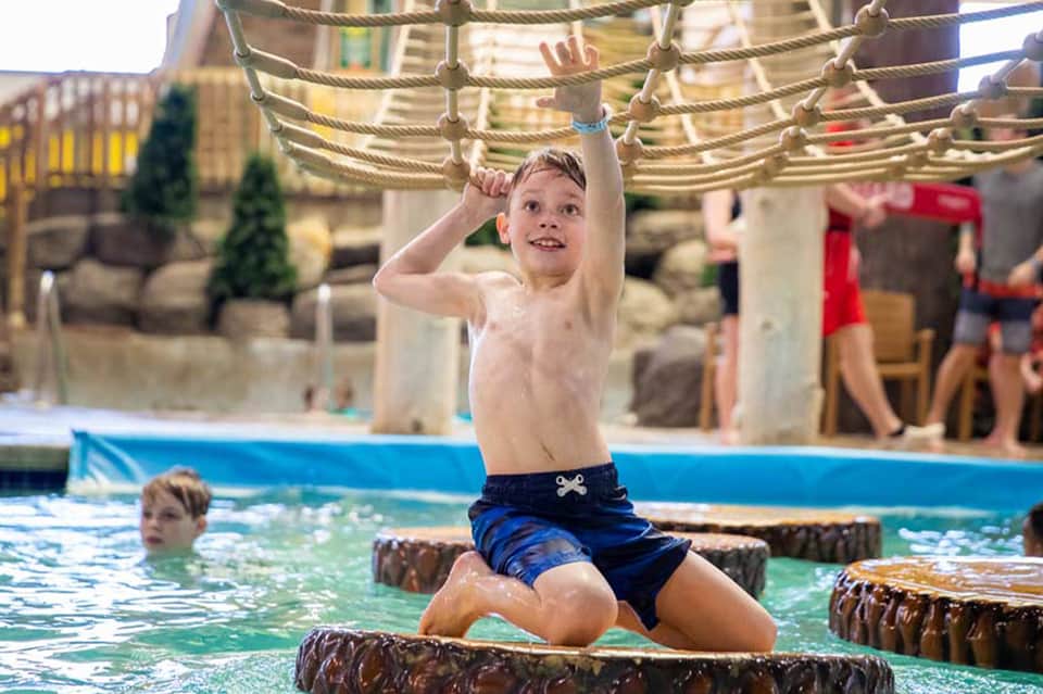 Young boy playing on rope obstacle course over shallow pool water at resort recreation area