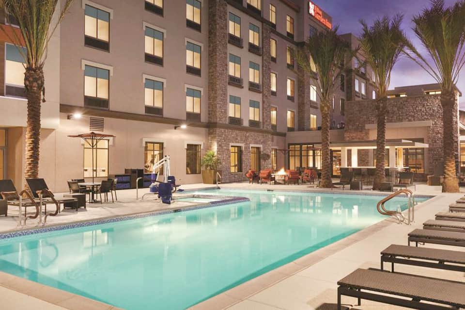 Hotel courtyard with illuminated rectangular pool, lounge chairs, palm trees, lit building facade, and evening atmosphere
