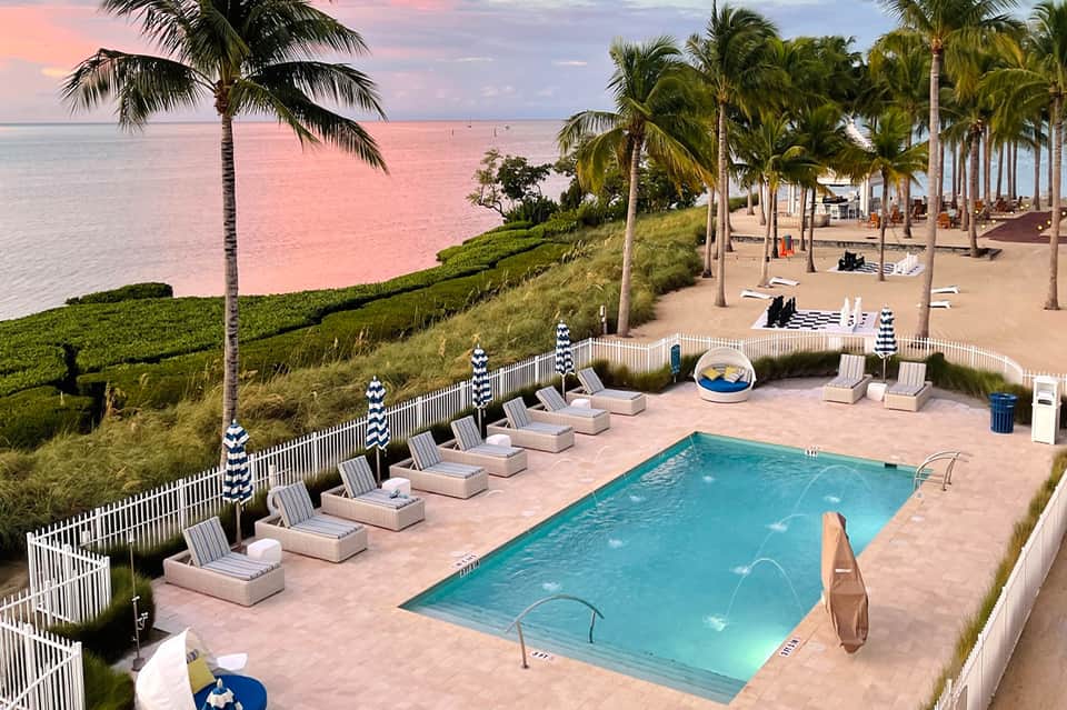 Beachfront pool with palm trees, striped umbrellas, lounge chairs, and ocean horizon at sunset