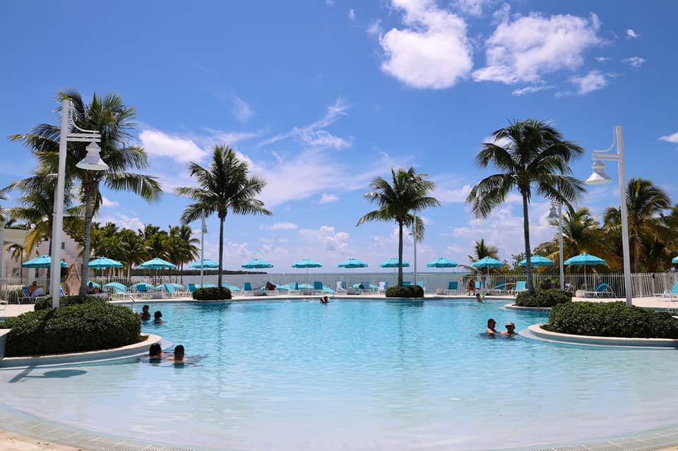 Resort pool with turquoise water, palm trees, white umbrellas, and guests swimming on sunny day