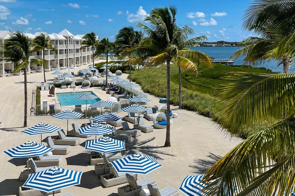 Beachfront resort with white building, blue umbrellas, palm trees, and waterfront view