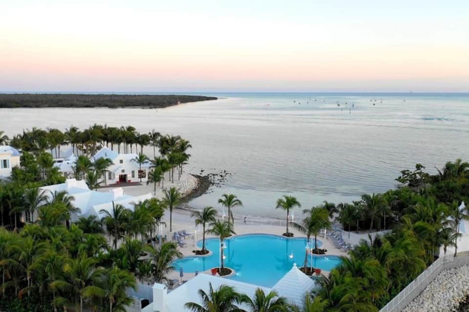 Luxury beachfront resort with white buildings, blue pool, palm trees, and calm ocean waters