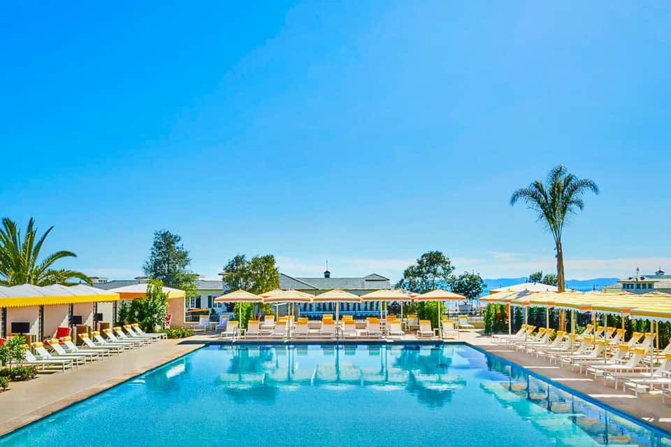 Resort swimming pool with yellow umbrellas, lounge chairs, palm trees, and clear blue sky.