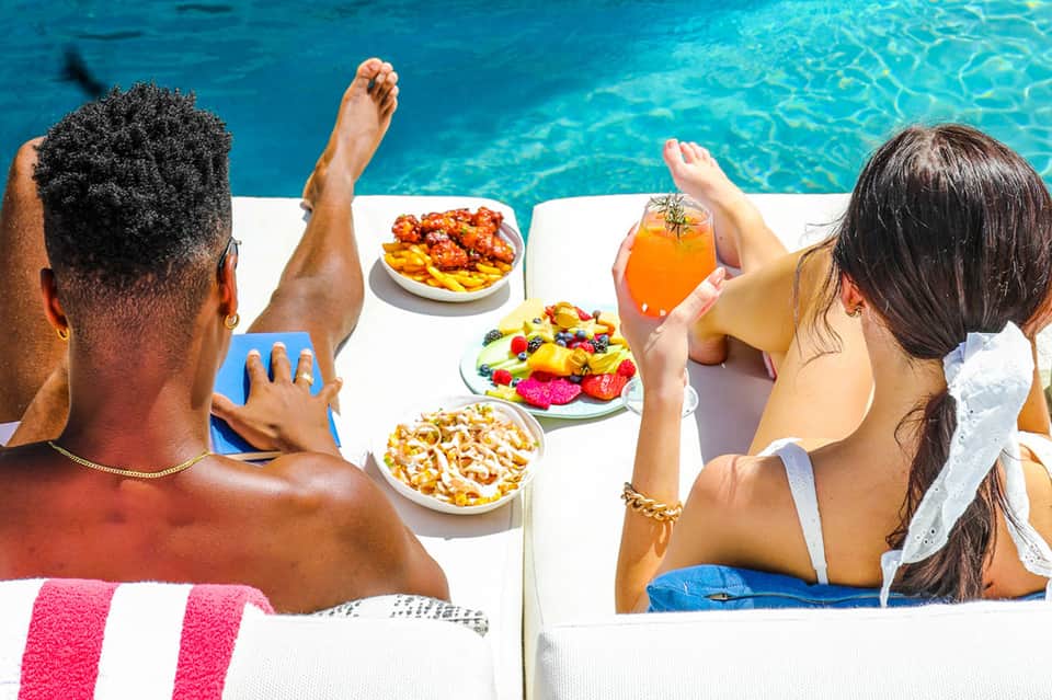 Guests relaxing poolside with colorful fruit platters and orange juice drinks by crystal blue water