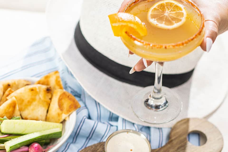 Woman in white hat holding citrus cocktail with orange slice, outdoor dining setting