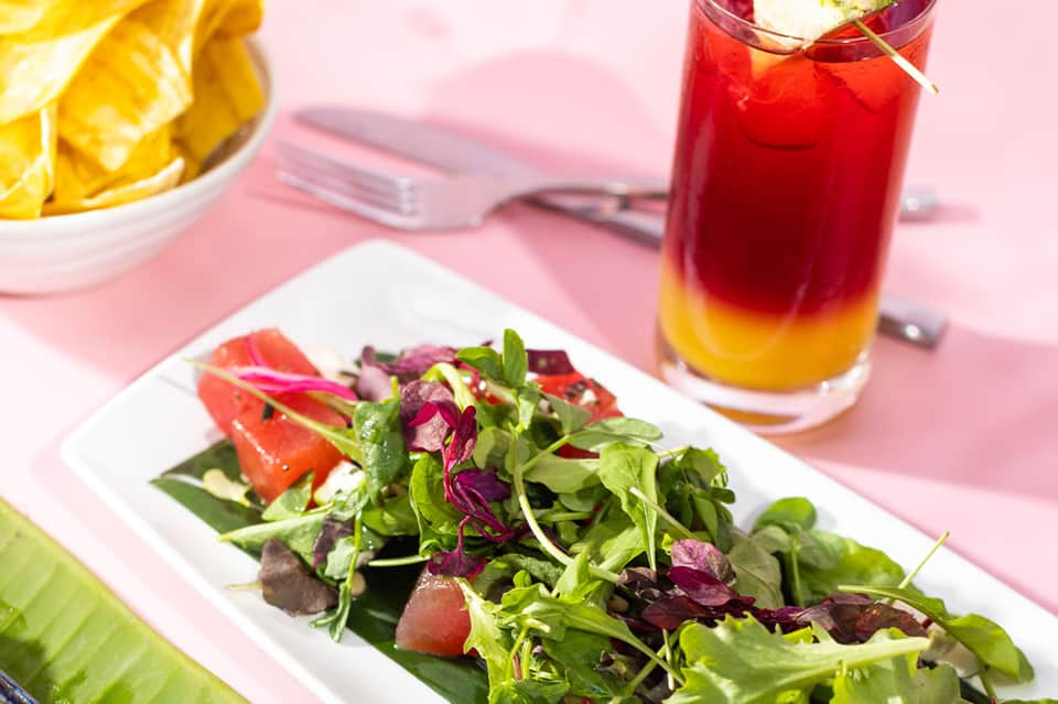 Colorful tropical meal with fresh salad, layered red and yellow cocktail, and citrus garnish on pink table