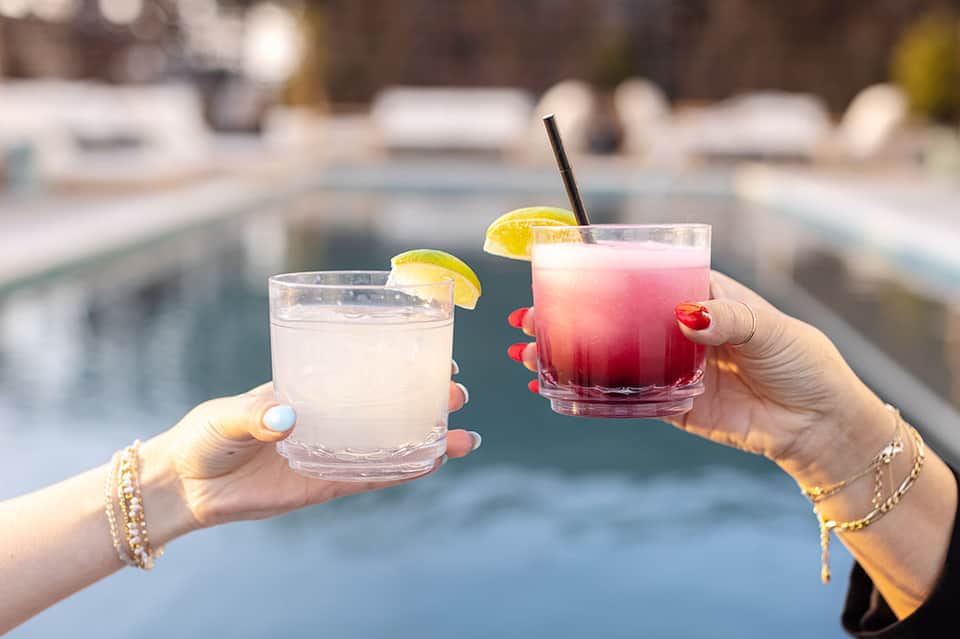 Two colorful cocktails with lime garnish held by pool with lounge area out of focus behind