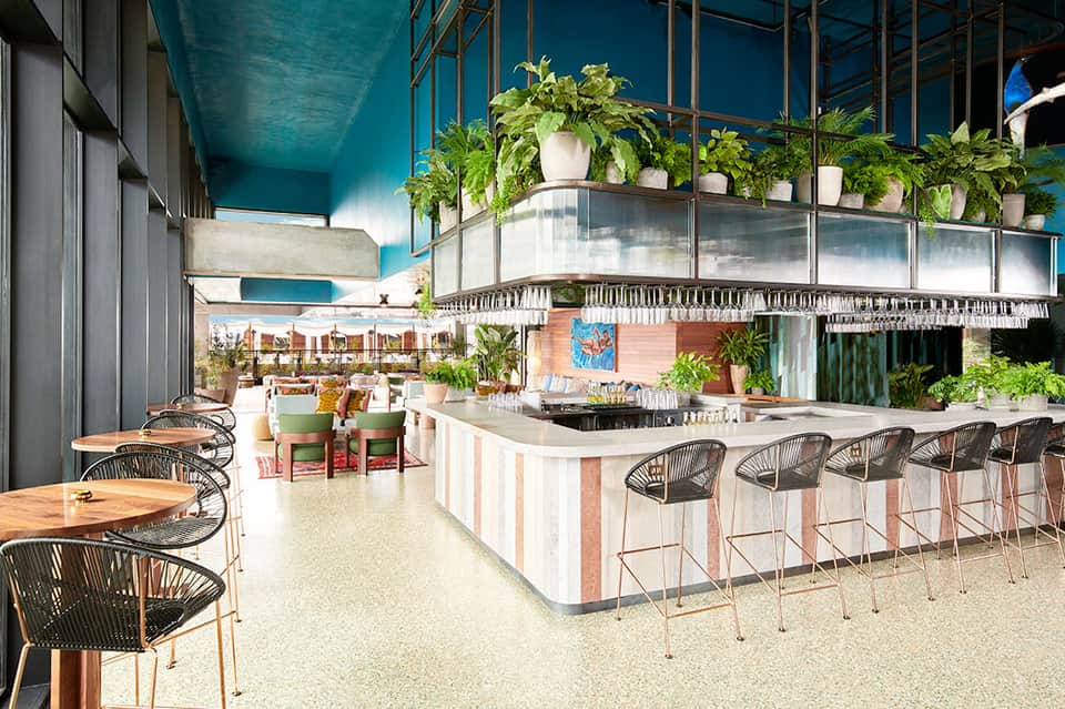 Modern hotel restaurant with turquoise ceiling, bar seating, and lush plant décor