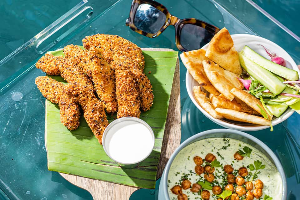 Breaded chicken strips and fries on green leaf plate by turquoise poolside table with sunglasses