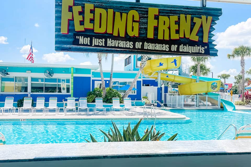 Bright turquoise resort pool with yellow waterslide, lounge chairs, palm trees, and feeding frenzy sign