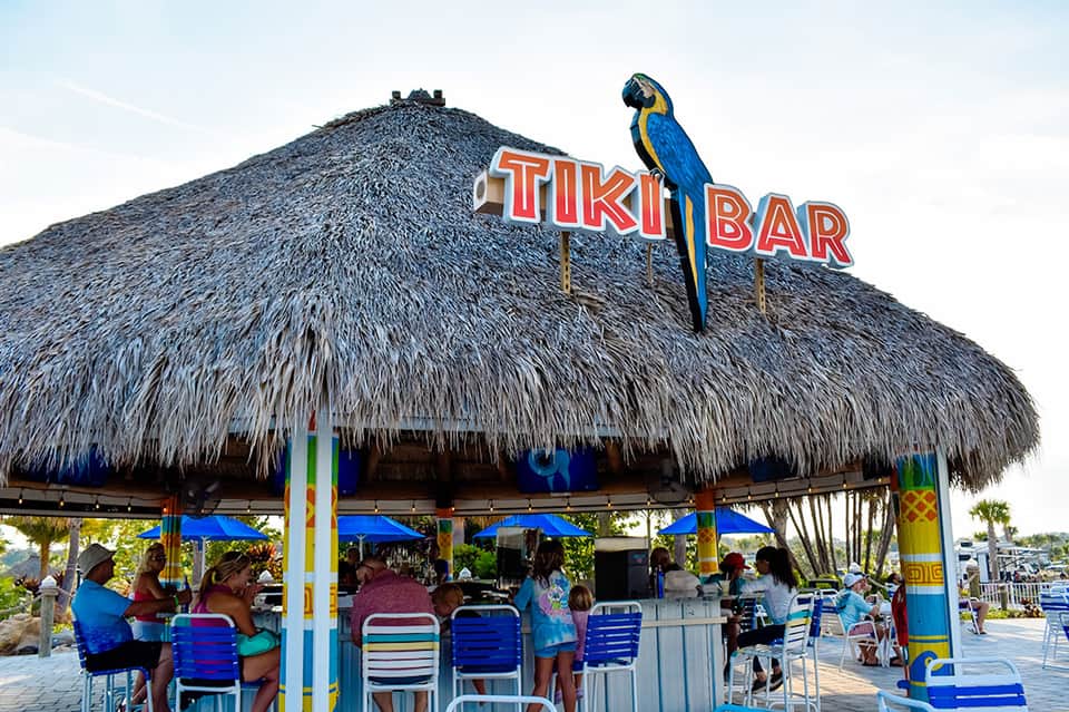 Colorful tiki bar with thatched roof, parrot sign, and beach patrons seated