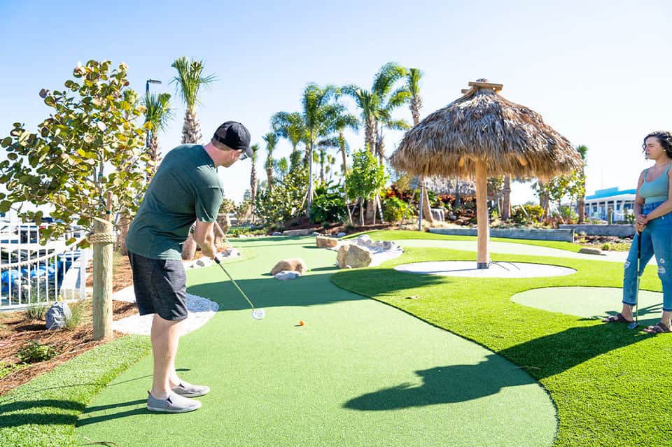 Family playing mini golf on green course with palm trees, tiki umbrella, and waterfront