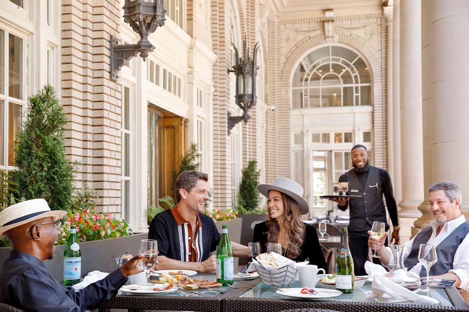 Upscale outdoor restaurant with guests dining at white tables on columned courtyard terrace with classical architecture