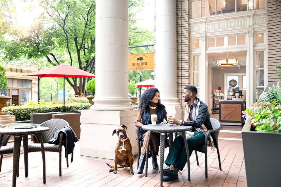 Urban courtyard seating area with red umbrellas, columns, and two people conversing with dog