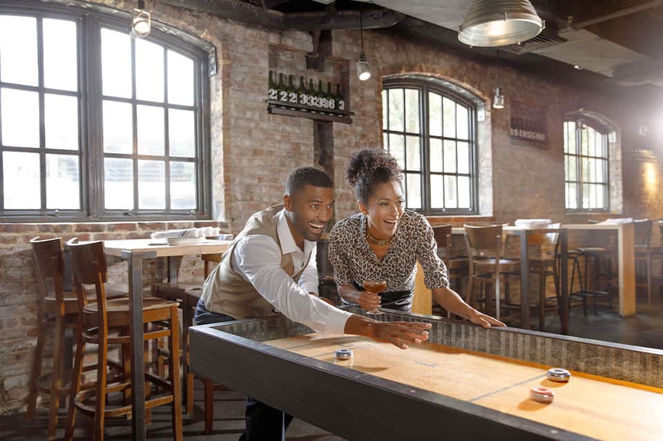 Couple playing shuffleboard in industrial-style bar with exposed brick, large windows, and wooden tables