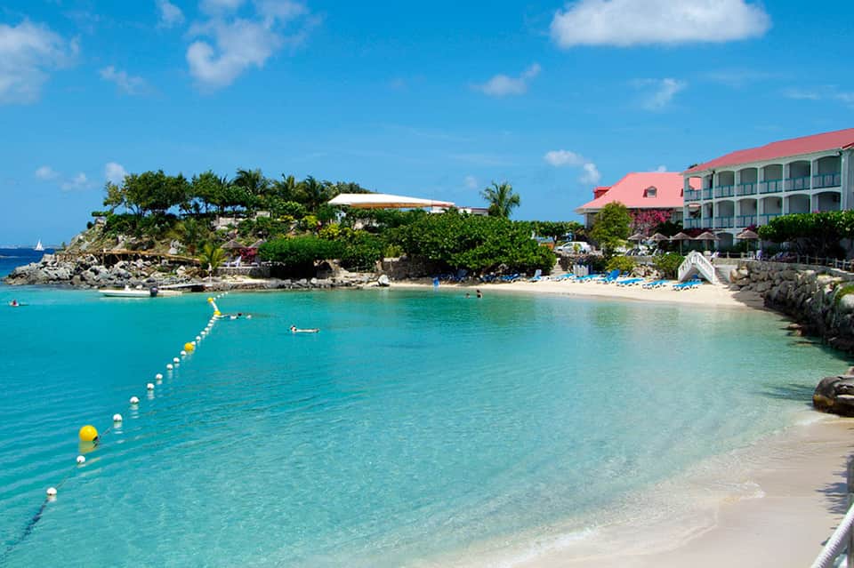 Pristine turquoise beach cove with white sand, resort buildings, and swimming area