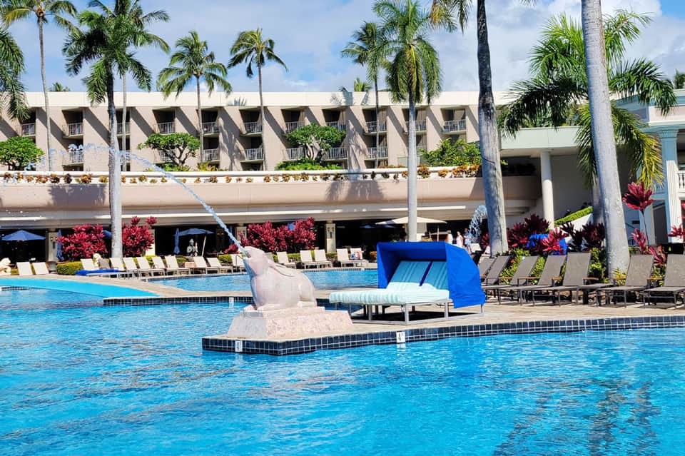 Resort pool with white statue sculpture, blue slide, palm trees, lounge chairs, and multi-story building